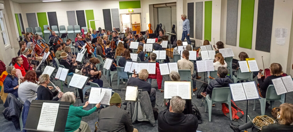 Photograph of symphony orchestra rehearsing, taken from behind the brass and second violins. Willem Van der Vis, a tall man with curly grey hair, is conducting. Several members of the orchestra are wearing puffer jackets and beanies.